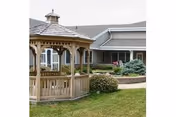 Outdoor view of a senior living facility featuring a wooden gazebo on a grassy lawn, with a building and landscaped bushes in the background under an overcast sky.