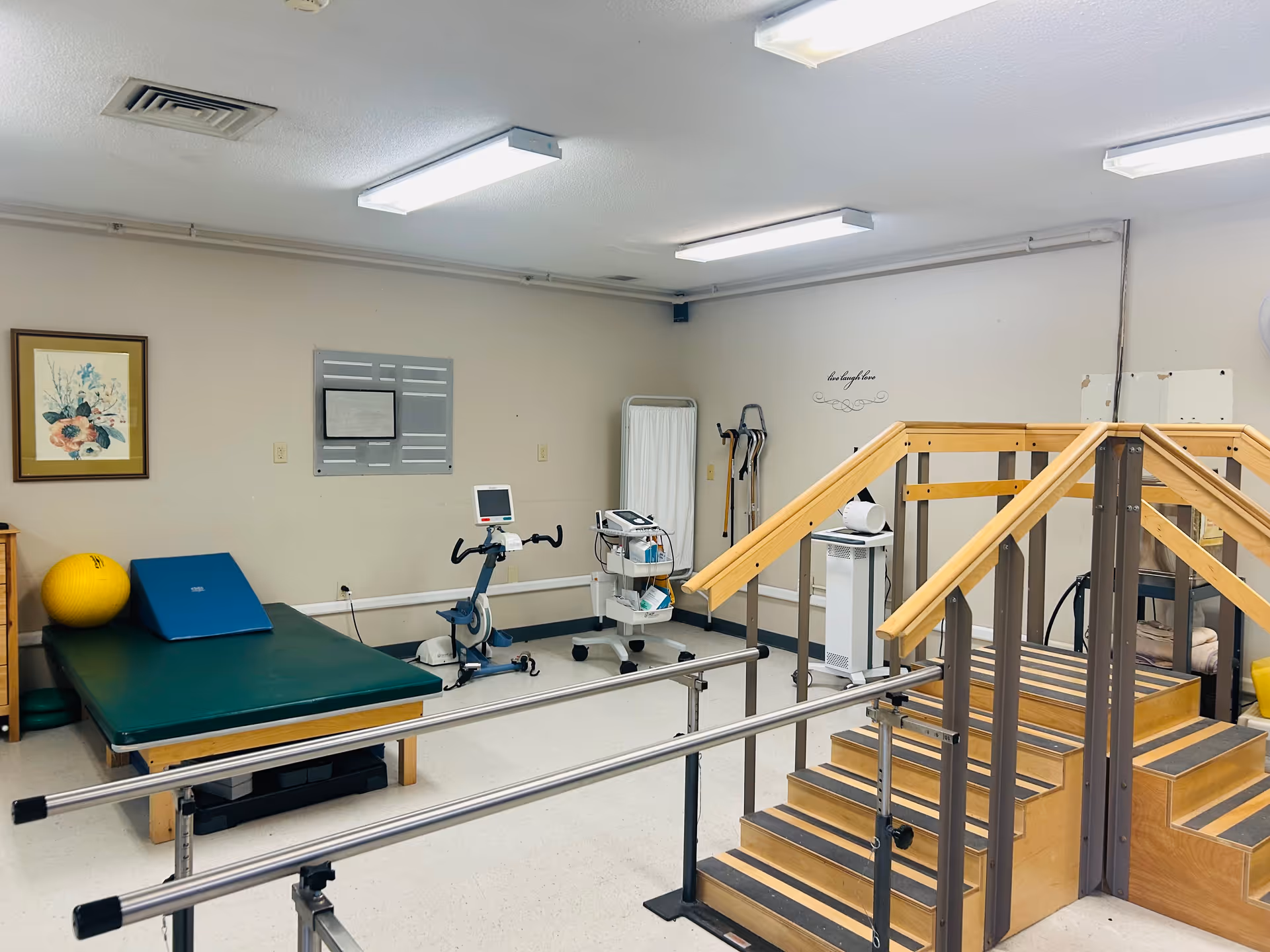 Therapy room with parallel bars and practice stairs, an exercise bike, treatment table, and other rehabilitation equipment.
