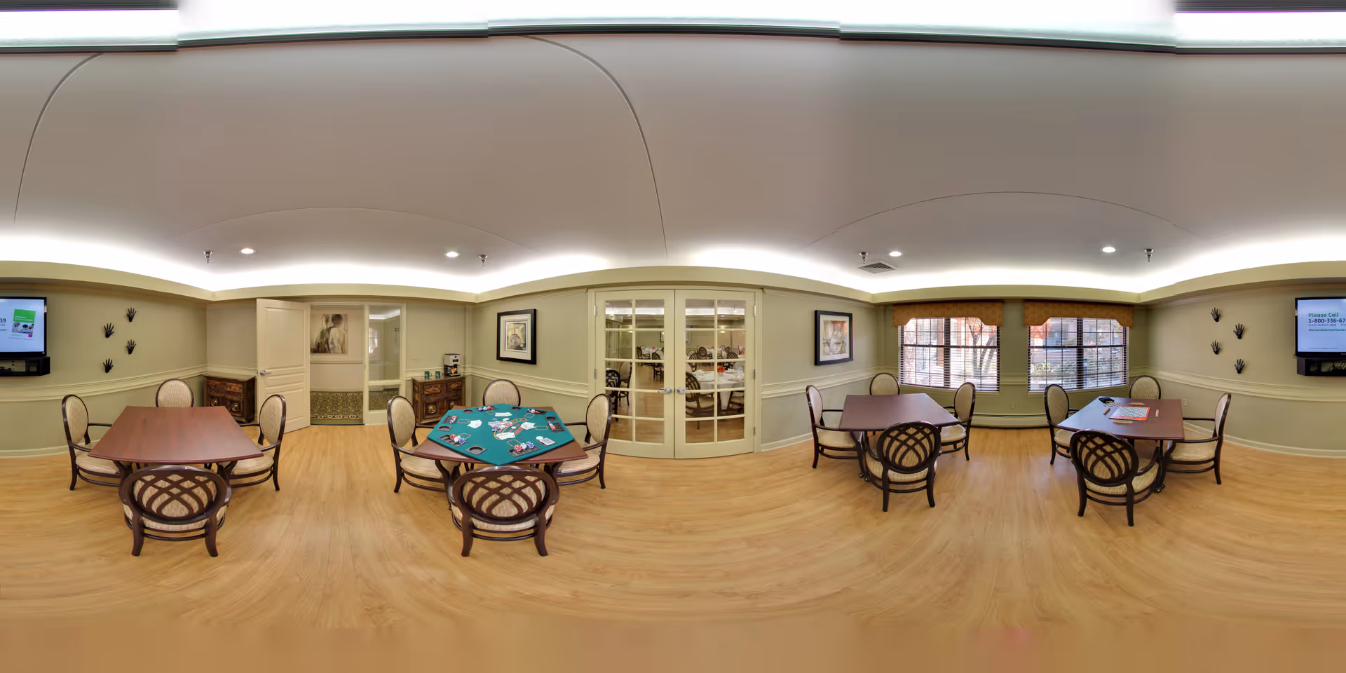 A spacious senior community activity room with wooden flooring, several tables and chairs arranged for group activities. One table has a green felt surface with playing cards and poker chips, suggesting a card game setup. The room has soft lighting with a curved ceiling design, wall-mounted televisions, framed artwork, and large windows letting in natural light. There are double glass doors leading to another room.