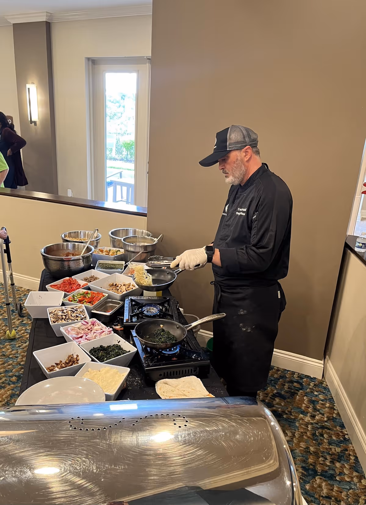 A chef wearing a black uniform and cap is cooking at a buffet-style setup with various bowls of fresh ingredients including vegetables, mushrooms, and cheese. The chef is preparing food on portable gas burners in an indoor setting with a window and beige walls.
