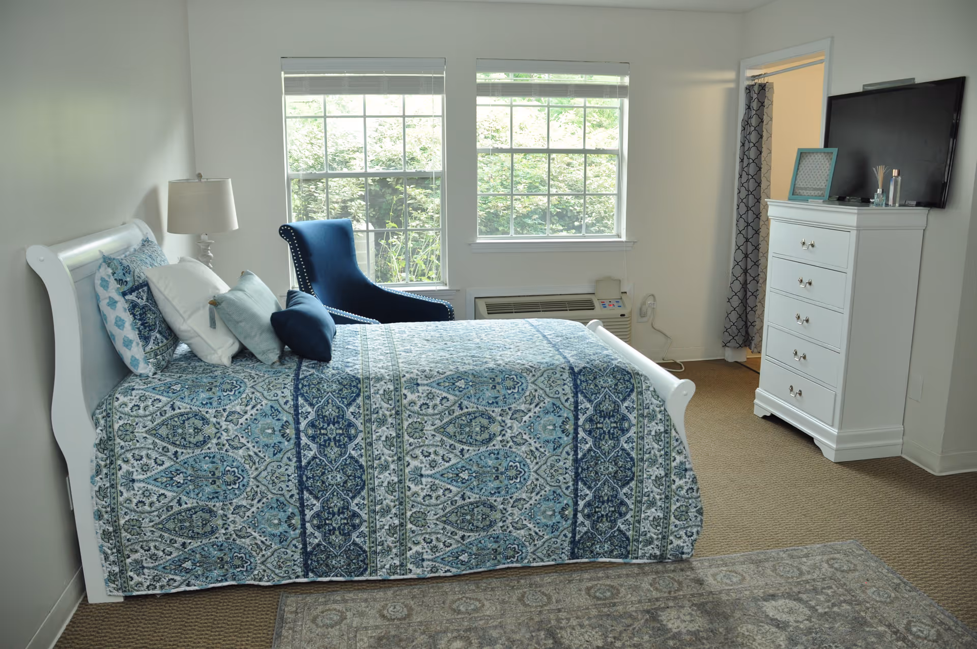 A bright bedroom with a white sleigh bed dressed in blue patterned bedding, a navy armchair by the windows, and a white dresser topped with a TV.