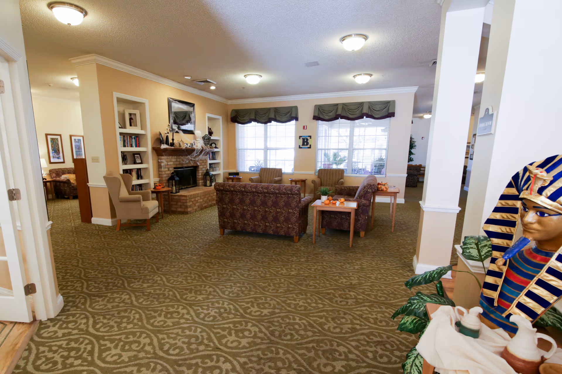 A cozy living room area in a senior living facility with patterned carpet, multiple armchairs and sofas arranged around a brick fireplace decorated with Halloween-themed items. Large windows with green valances allow natural light to fill the room. A decorative Egyptian pharaoh bust is visible on the right side near some plants and ceramic pitchers.