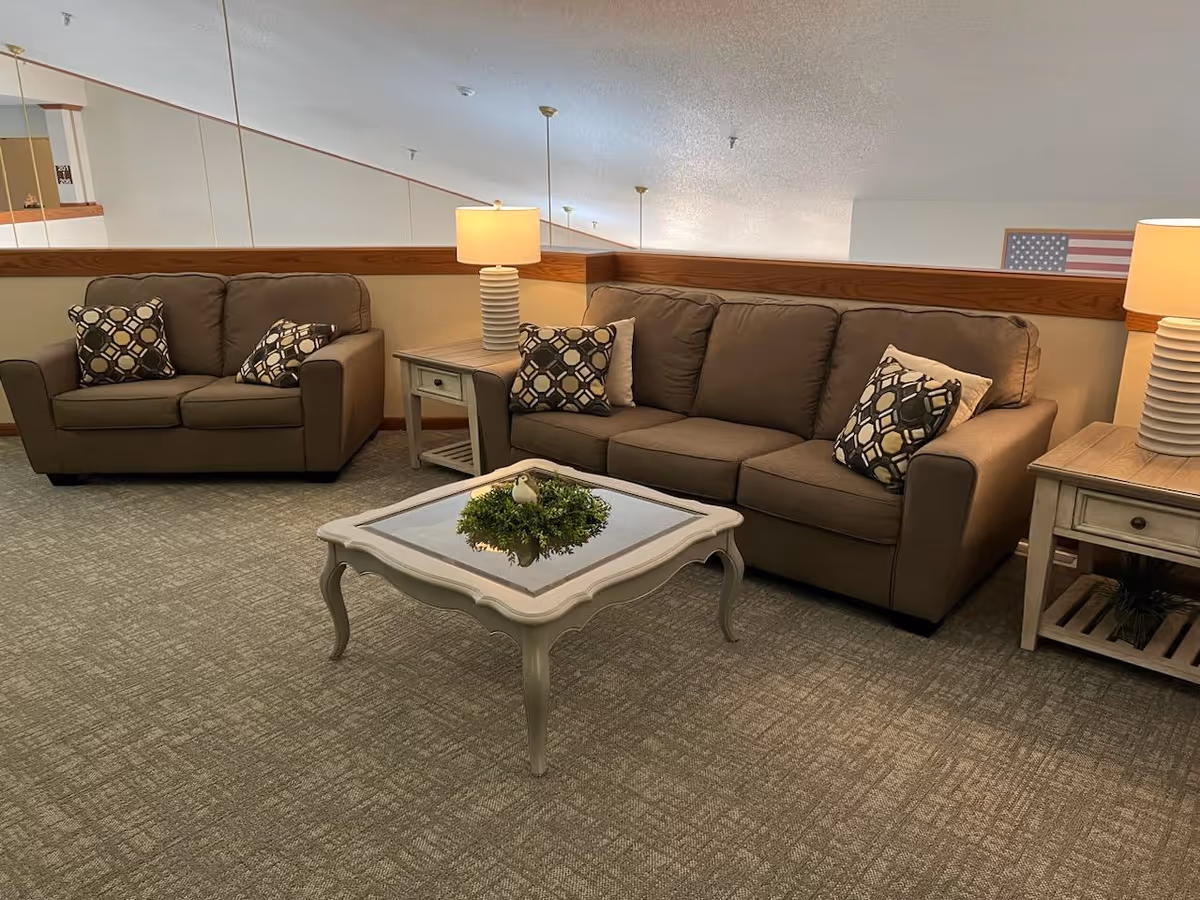 A cozy living room area with two brown sofas adorned with patterned throw pillows. Between the sofas is a wooden side table with a white lamp. In front of the sofas is a white coffee table with a glass top and a small green plant centerpiece. The room has carpeted flooring and a wooden railing along the back wall.