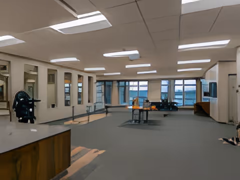 A spacious interior room with large windows along one wall, carpeted floor, and ceiling lights. There are tables and chairs arranged in the middle and some furniture along the walls. The room appears to be a common area or lounge in a senior living facility.