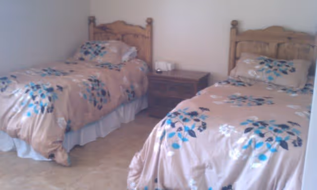 A bedroom with two single beds featuring matching floral-patterned bedspreads in shades of beige, blue, and brown. Between the beds is a wooden nightstand with a white tissue box on top. The room has a tiled floor and plain white walls.