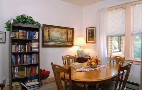 A wood dining table with chairs, bookshelf, framed art, and windows in a bright dining area.