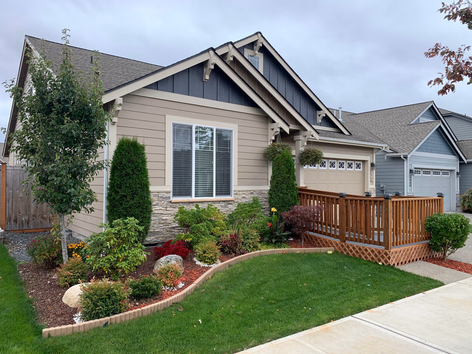 Exterior view of a single-story residential building with beige siding and stone accents, featuring a wooden ramp leading to the front door. The front yard is landscaped with green grass, various shrubs, small trees, and flowers under a cloudy sky.