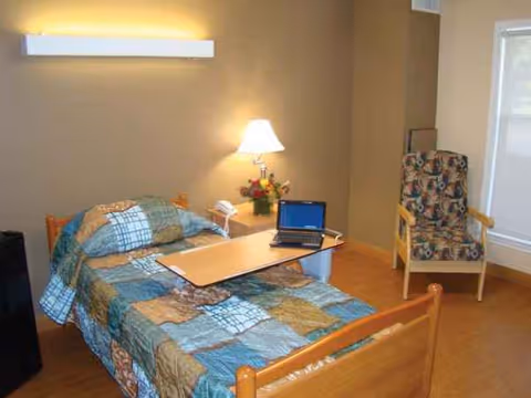 A furnished senior living facility bedroom with a bed, quilt, chair, lamp, and laptop.