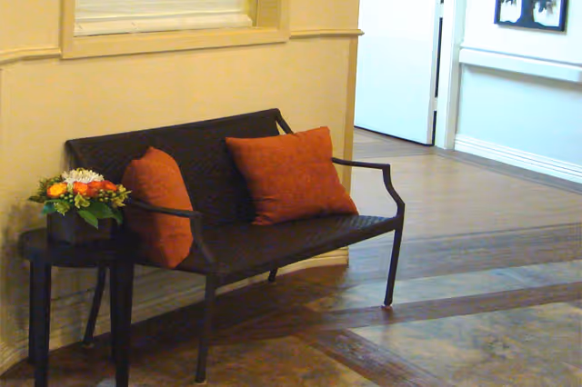 Metal bench with two orange pillows and a small side table holding a floral arrangement in a hallway interior.