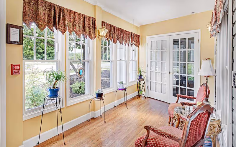 A bright sunroom with large windows covered by patterned valances, several potted plants on metal stands along the window ledge, two red upholstered chairs with wooden frames, a small glass-top table, a floor lamp, and French doors leading to another room.