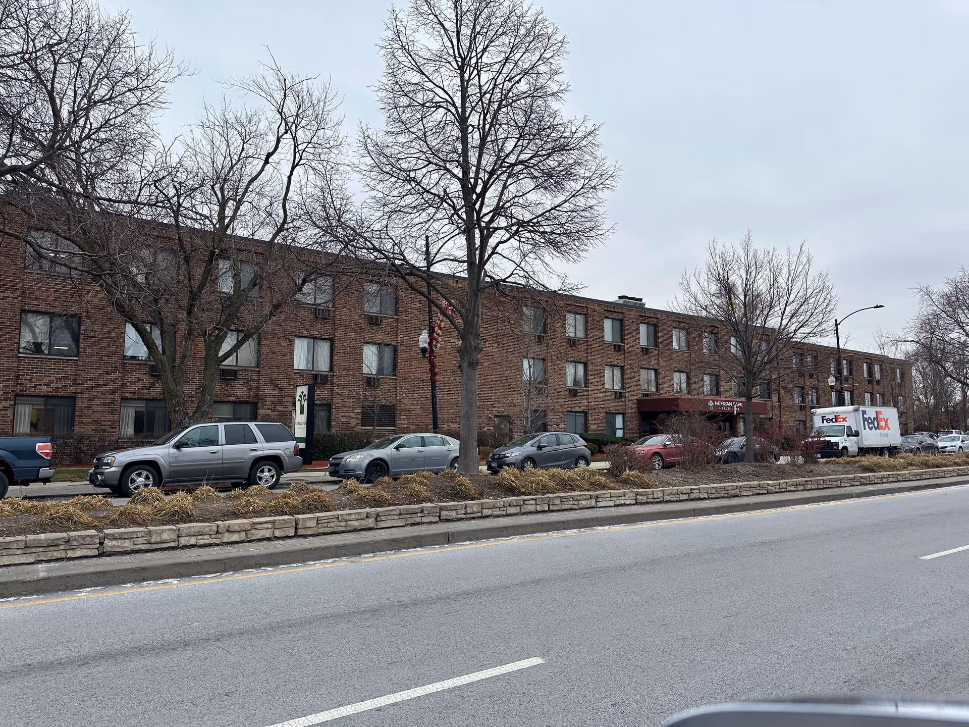 Three-story brick apartment-style building with parked cars and bare trees along the street on an overcast day.