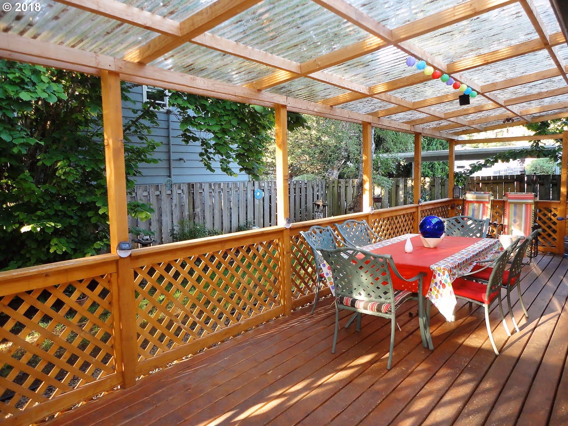 Covered outdoor wooden deck with a dining table set for six, featuring a red tablecloth and a decorative blue glass orb centerpiece. The deck has lattice-style wooden railings and a transparent corrugated roof. Colorful string lights hang from the ceiling. Surrounding the deck are green trees and a wooden fence.