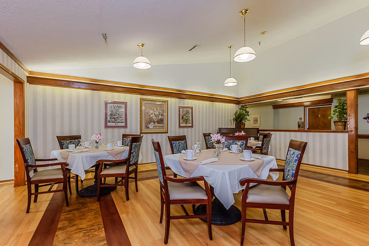 A dining room with round tables set with tablecloths and place settings, surrounded by wooden chairs under pendant lights.