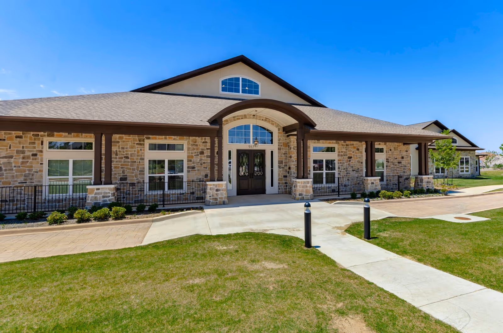 Exterior front view of a single-story building with stone and beige siding, large windows, and a covered entrance with double doors. The building is surrounded by a well-maintained lawn and a paved walkway leading to the entrance under a clear blue sky.