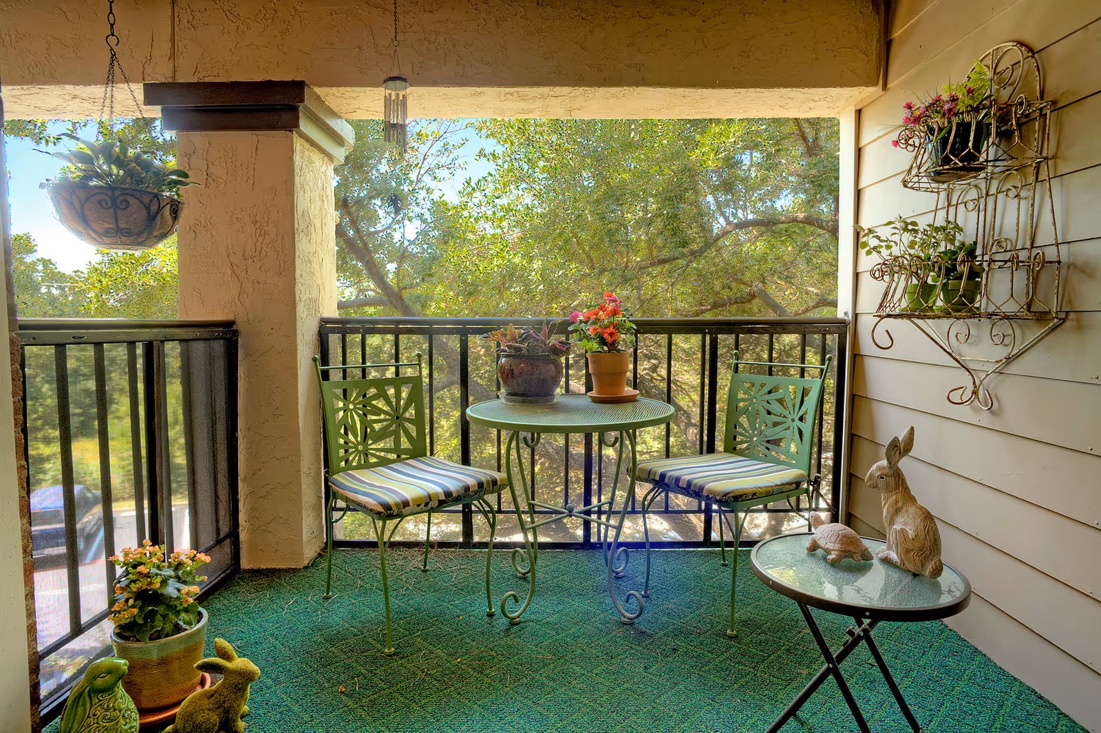 A cozy green-carpeted balcony with a small round table, two chairs with striped cushions, potted plants, and decorative rabbit statues overlooking trees.