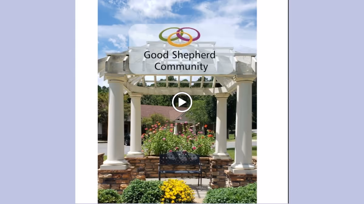 Outdoor pergola structure with white columns and a stone base, surrounded by green bushes and yellow flowers, with a black metal bench in the center. In the background, there is a building with a brown roof and trees under a partly cloudy blue sky.