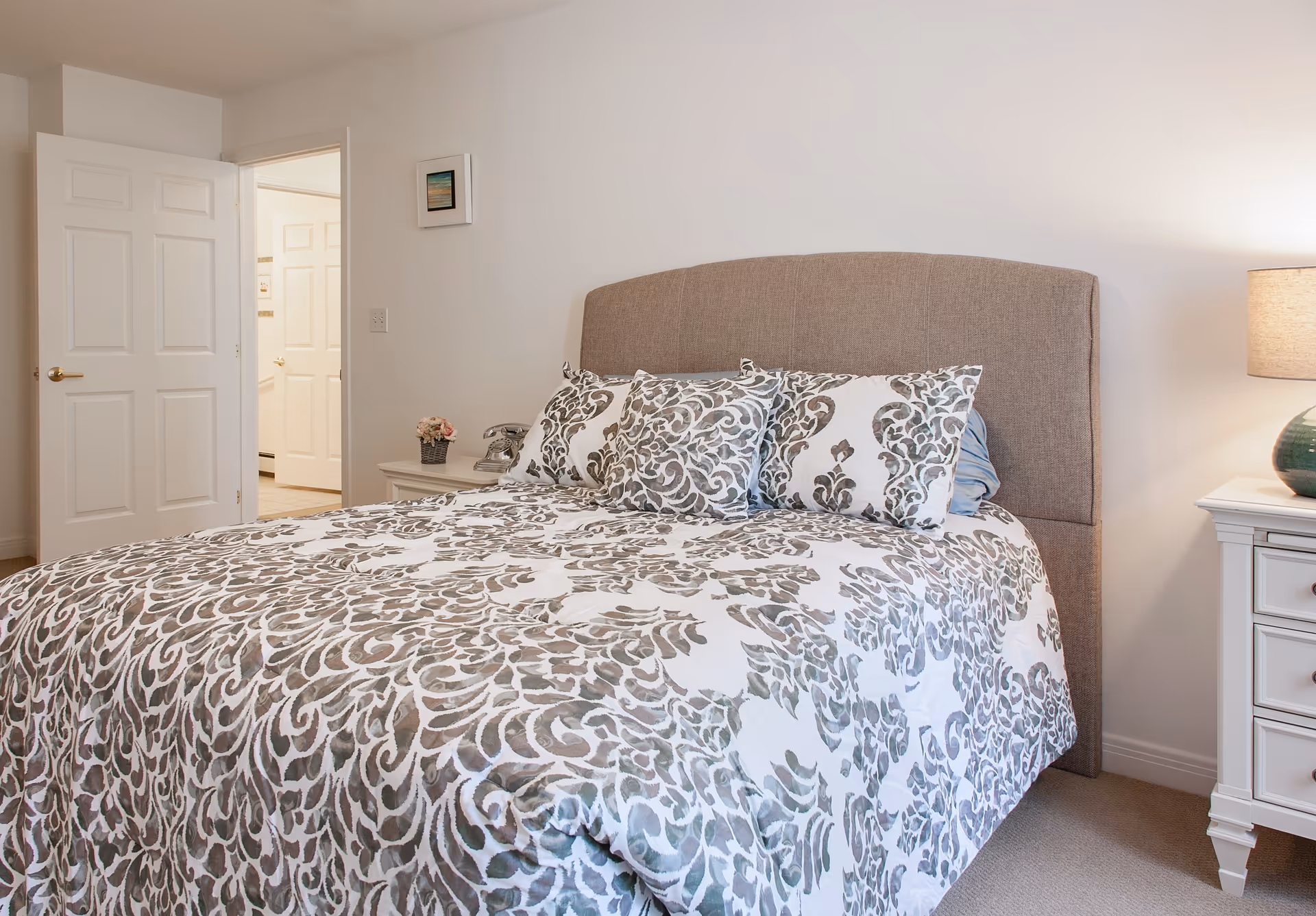 A bedroom with a large bed featuring a beige upholstered headboard and a white and gray patterned bedspread with matching pillows. There are two white nightstands on either side of the bed, one with a small flower arrangement and an old-fashioned telephone, and the other with a green lamp. The room has light-colored walls and carpet, with an open door leading to another room.