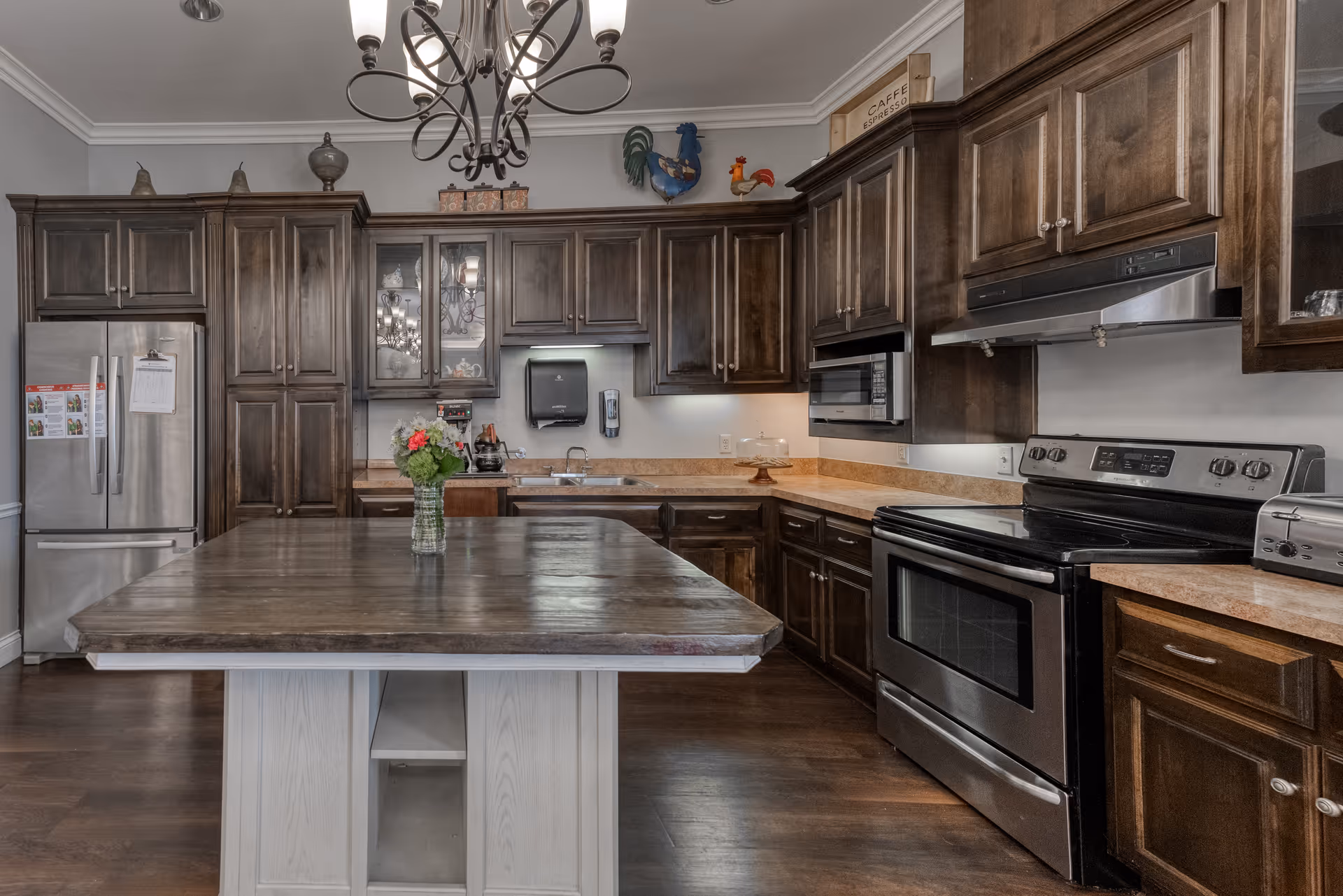 A modern kitchen with dark wooden cabinets, stainless steel refrigerator, oven, microwave, and a large wooden island in the center with a vase of flowers. The kitchen has a chandelier hanging from the ceiling and various decorative items on top of the cabinets.