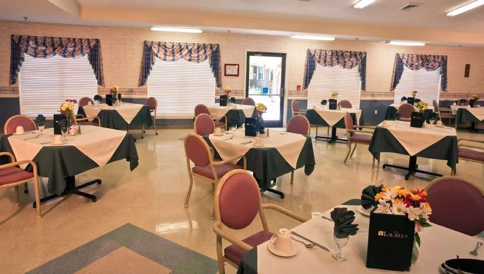 Dining room with multiple tables covered with white and green tablecloths, each set with utensils, napkins, cups, and flower centerpieces. The room has several windows with patterned valances and a door leading outside.