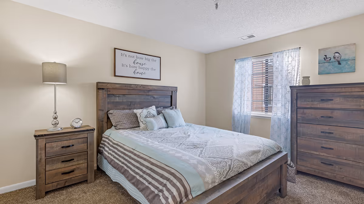 A cozy bedroom with a wooden bed frame and matching wooden nightstand and dresser. The bed is made with patterned bedding and several pillows. A lamp and a small clock sit on the nightstand. A window with light blue patterned curtains lets in natural light. Wall decor includes a framed quote above the bed and a painting of birds on the wall near the dresser.
