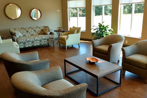 A bright common room with a sofa, several armchairs, a central coffee table, and large windows letting in daylight.