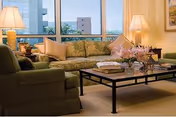 A cozy living room with a green sofa and armchair, a black metal coffee table with books and a flower arrangement, two table lamps on side tables, and large windows showing a cityscape outside.
