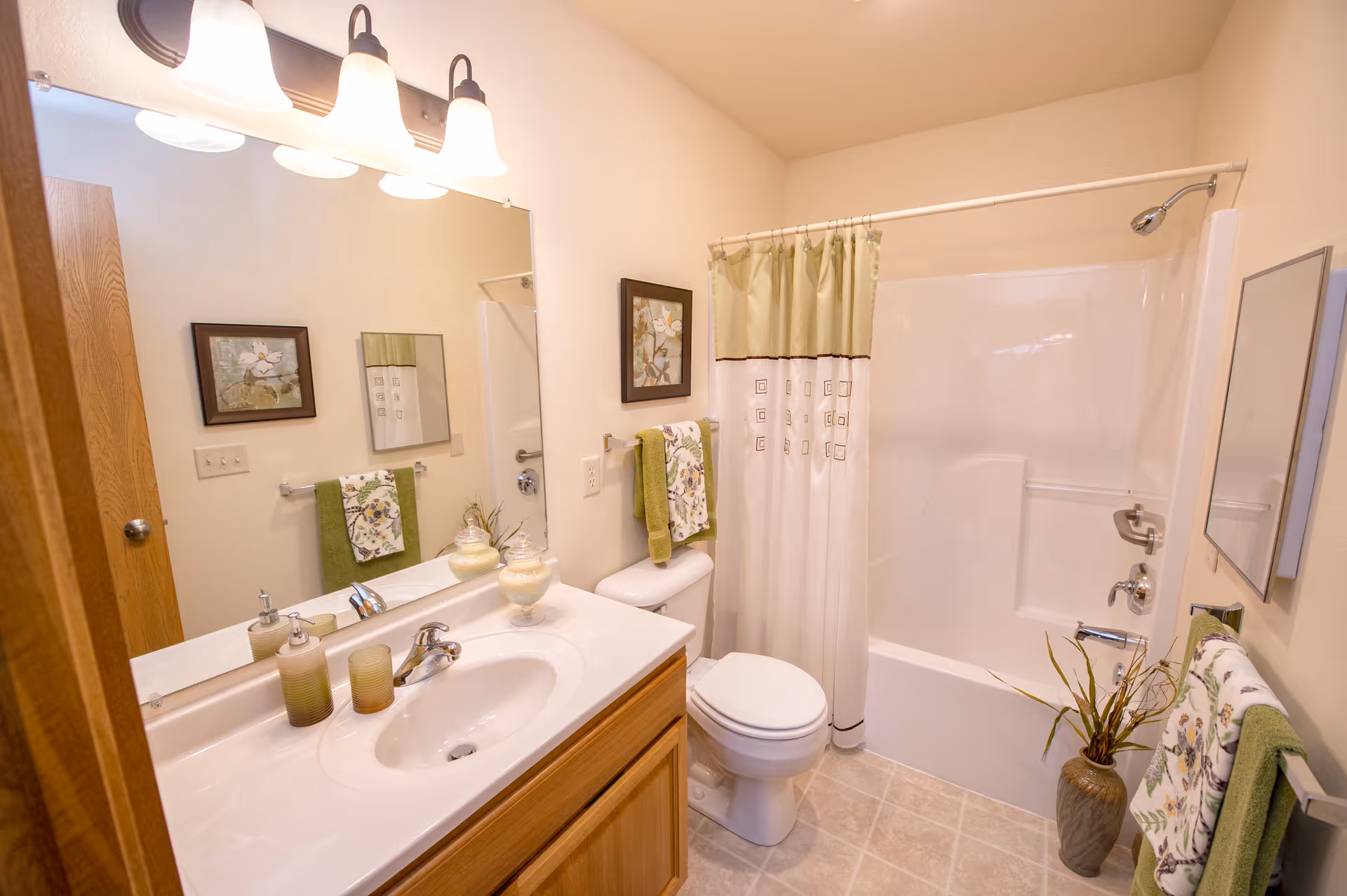 A clean and well-lit bathroom featuring a white sink with a wooden cabinet, a large mirror with three overhead lights, a toilet, and a bathtub with a shower curtain. The bathroom is decorated with green and floral towels, framed artwork on the walls, and a vase with plants on the floor near the bathtub.