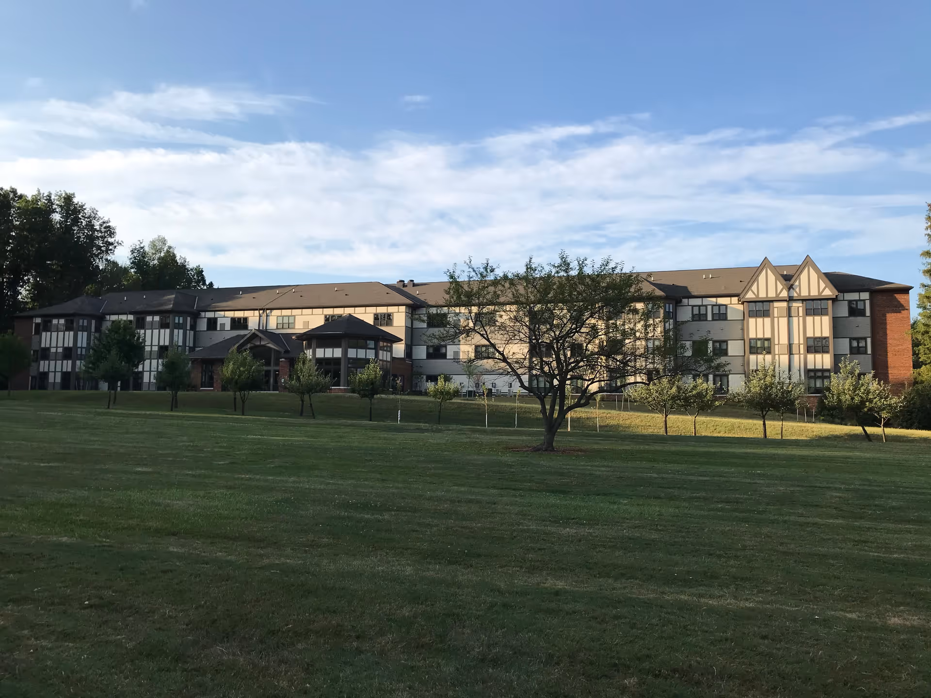A large multi-story assisted living facility building with a mix of brick and light-colored paneling, surrounded by a grassy lawn with several small trees and a partly cloudy sky above.