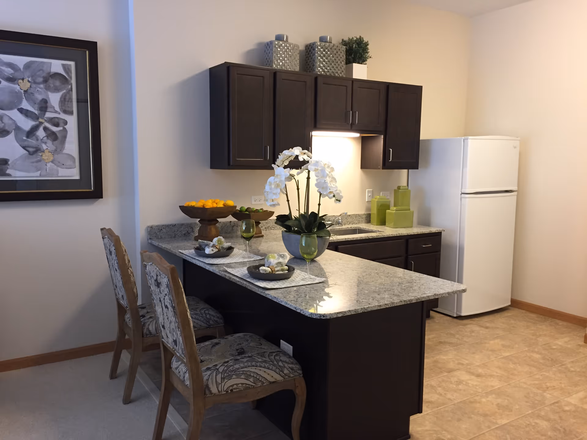Small kitchen with a granite breakfast bar, two upholstered bar stools, dark cabinets, a white refrigerator and decorative centerpiece.