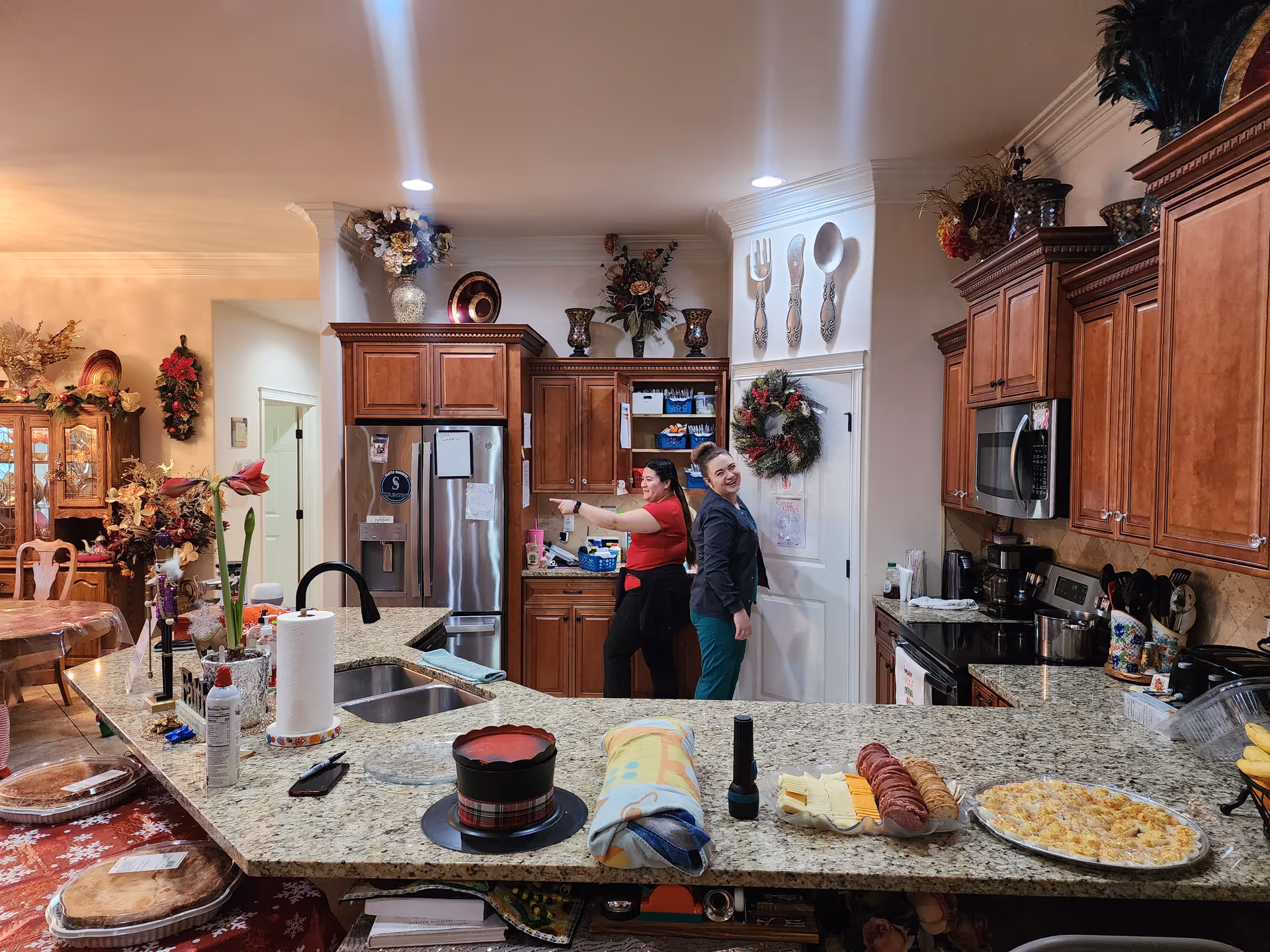 A warm, decorated kitchen with granite countertops and wooden cabinets. Two women are standing near the refrigerator and pantry, smiling and posing playfully. The kitchen is adorned with festive wreaths, floral arrangements, and large decorative utensils on the wall. Various food items and kitchen supplies are placed on the counter, including a pie, paper towels, and a flashlight.
