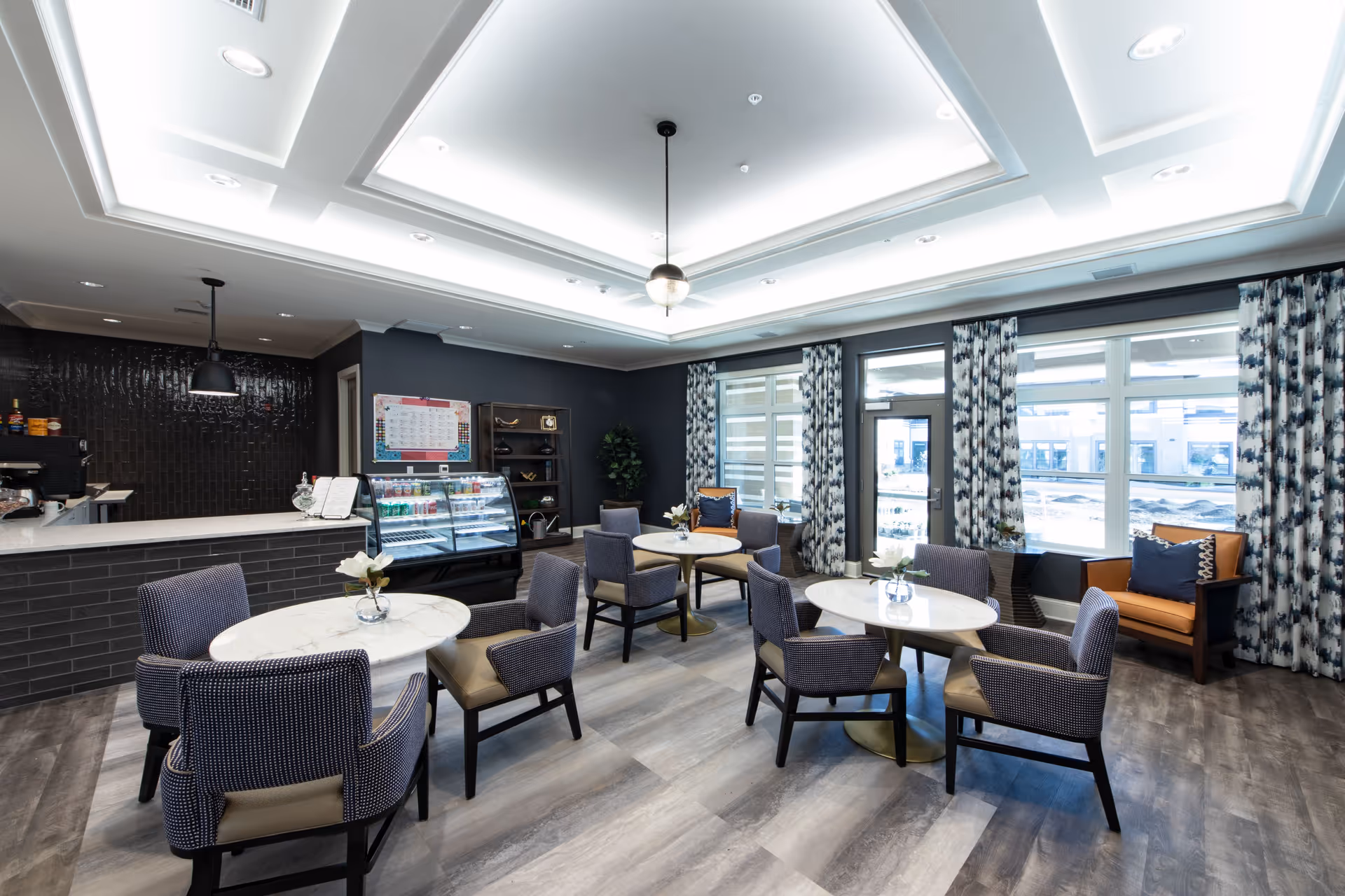 Bright modern common dining area with round marble-topped tables, upholstered chairs, a service counter with display case, and large windows.