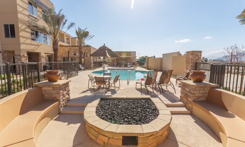 Outdoor courtyard featuring a swimming pool with lounge chairs and tables under an umbrella, a central stone fire pit, and surrounding building and palm trees.