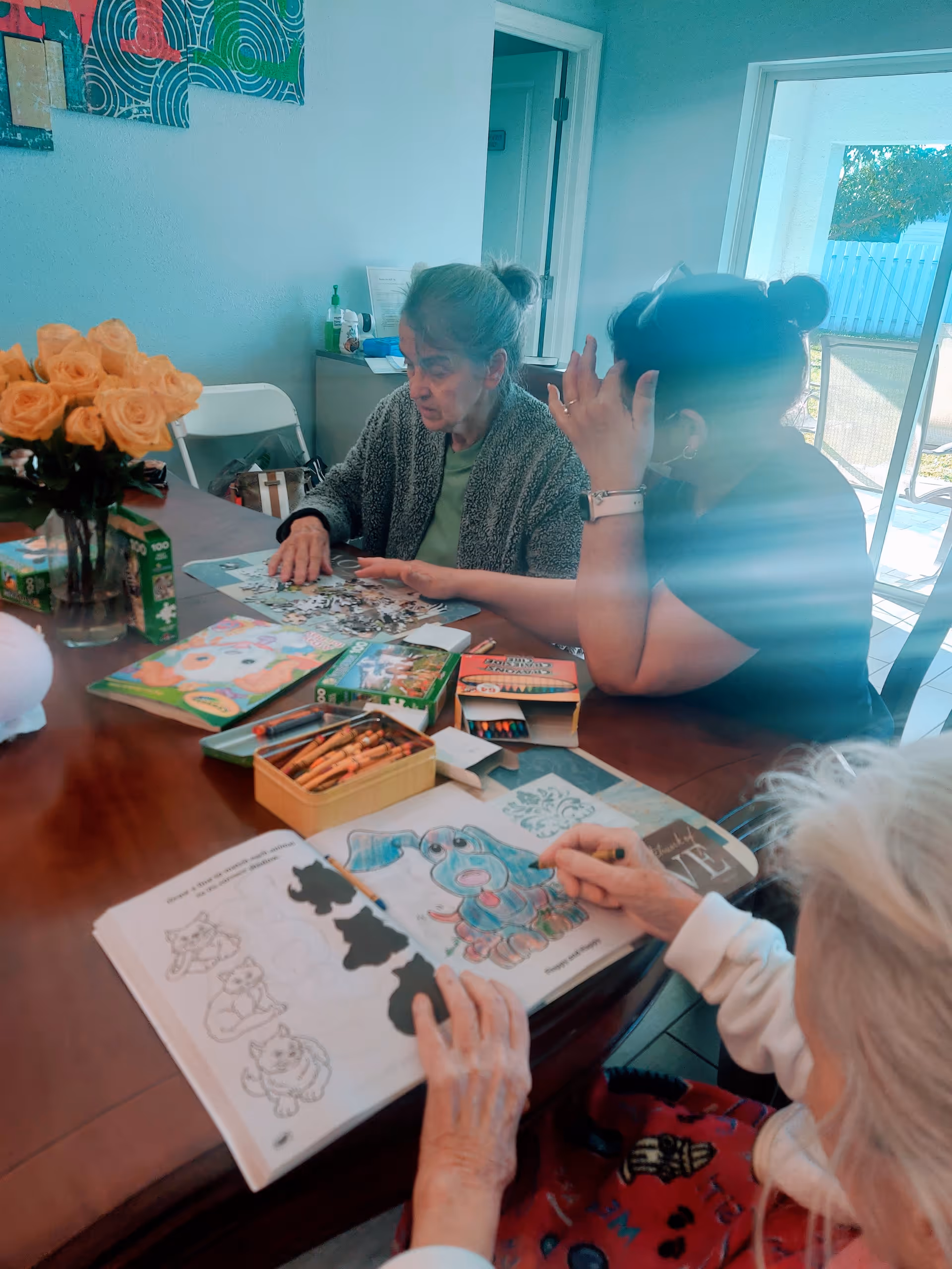 Three elderly women sitting around a table engaged in activities including coloring and working on a puzzle. The table has coloring books, crayons, and a vase with yellow roses. The room has light blue walls and a door leading outside with sunlight coming through.
