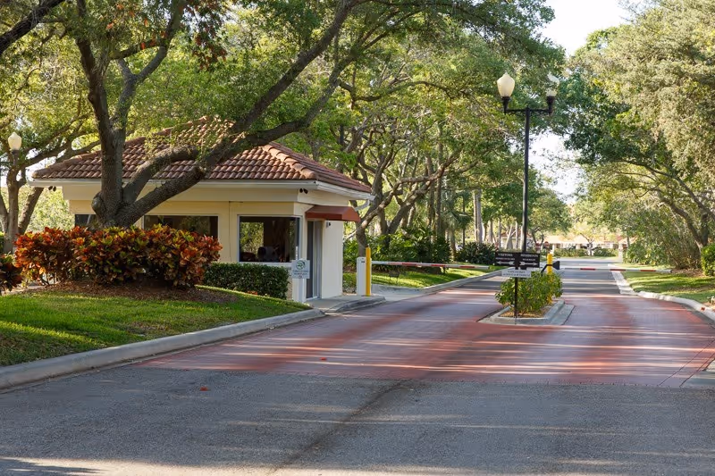 Tree-lined entrance road with a small guardhouse and security gate at a residential community.