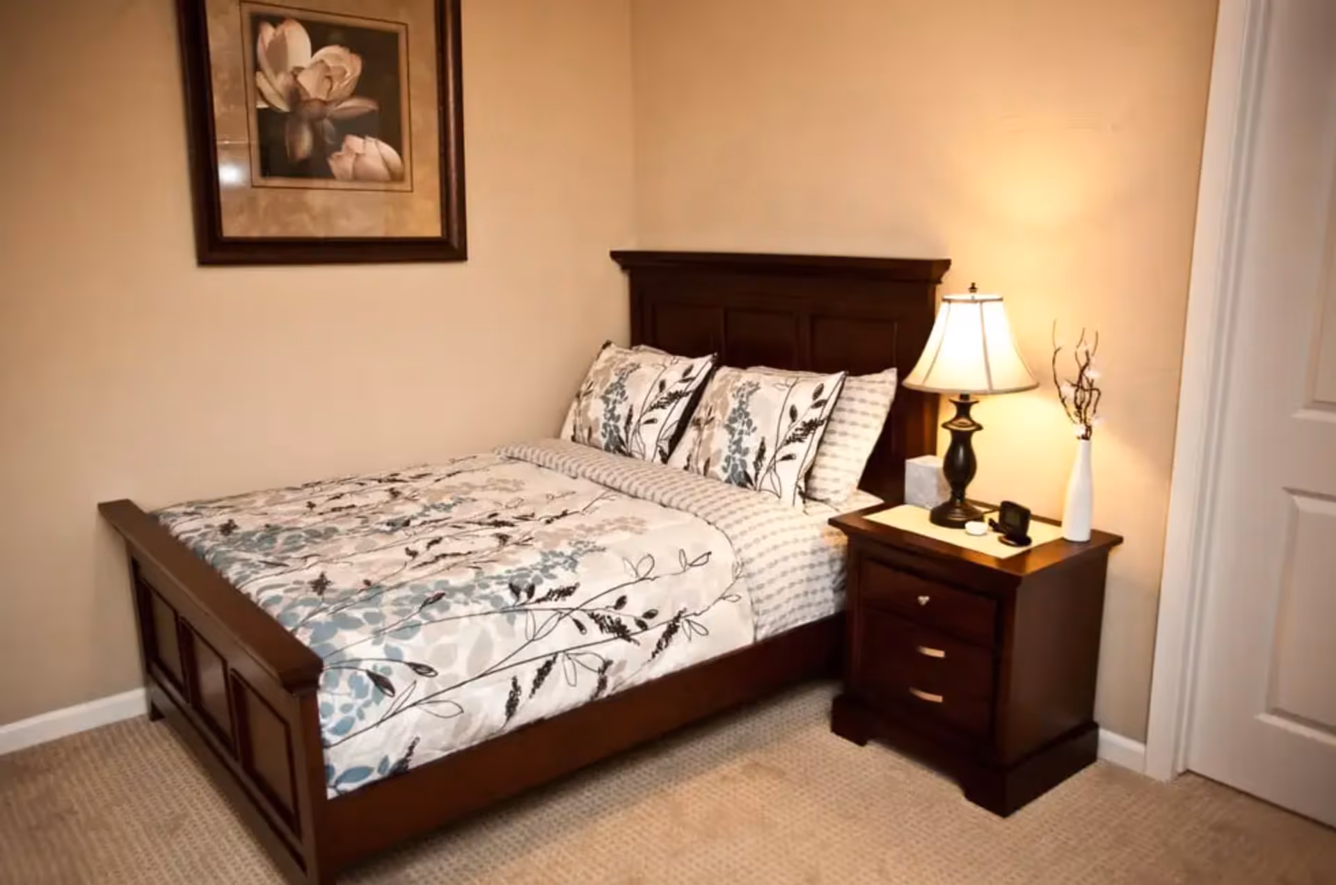 A bedroom with a wooden bed frame and a floral patterned bedspread and pillows. Next to the bed is a wooden nightstand with a lamp, a small clock, a tissue box, and a white vase with decorative branches. A framed floral painting hangs on the beige wall above the bed.