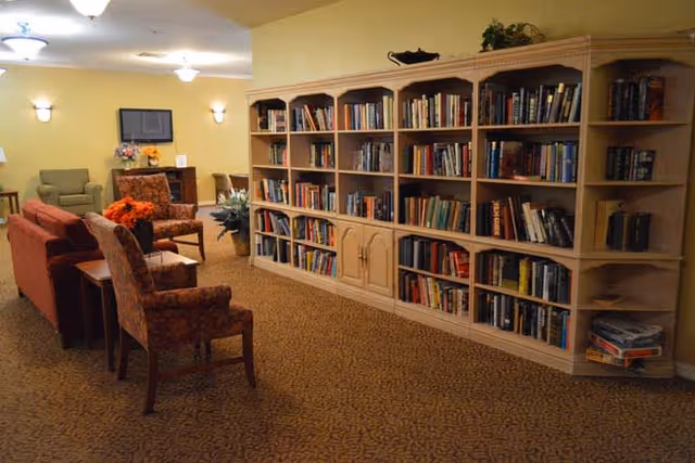 A cozy library or reading area in a senior living facility with a large wooden bookshelf filled with books along one wall. The room has several upholstered chairs and a sofa arranged for comfortable seating. The walls are painted yellow, and there are wall-mounted lights and ceiling lights providing warm illumination. A small table with a flower arrangement is also visible.
