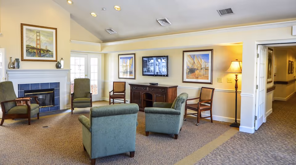 A cozy senior living community lounge with four green upholstered armchairs and two wooden chairs arranged around a fireplace and a TV mounted on the wall. The room features beige walls with framed artwork, a standing lamp, and carpeted flooring. French doors and a hallway are visible in the background.