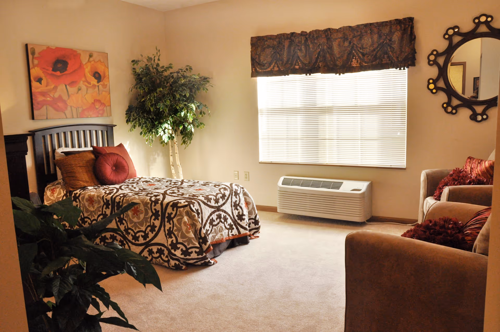 A cozy bedroom with a single bed covered in a patterned bedspread and decorative pillows. There is a large window with blinds and a valance, a potted plant in the corner, a cushioned armchair with pillows, a decorative wall mirror, and a floral painting on the wall.