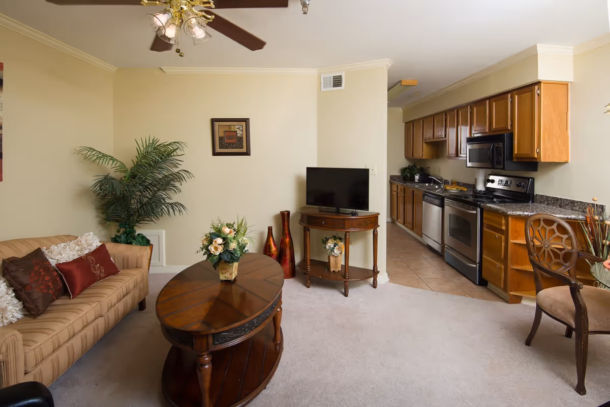 A cozy living room area with a beige striped sofa adorned with decorative pillows, a wooden coffee table with a floral arrangement, and a small wooden TV stand holding a flat-screen television. To the right, there is a kitchen with wooden cabinets, stainless steel appliances including a stove, microwave, and dishwasher, and a tiled floor. A ceiling fan with lights is visible above, and a potted plant and framed artwork decorate the walls.