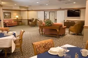 A spacious common area in a senior living facility with multiple seating arrangements including armchairs and sofas. There are tables set with white and blue tablecloths, cups, and plates, suggesting a dining or social area. The room has carpeted floors, neutral-colored walls, framed artwork, and a flat-screen TV mounted on the wall.
