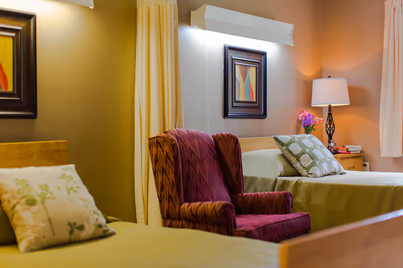 Two single beds in a warm-colored senior bedroom with a red upholstered armchair, bedside lamp, pillows, and framed art.