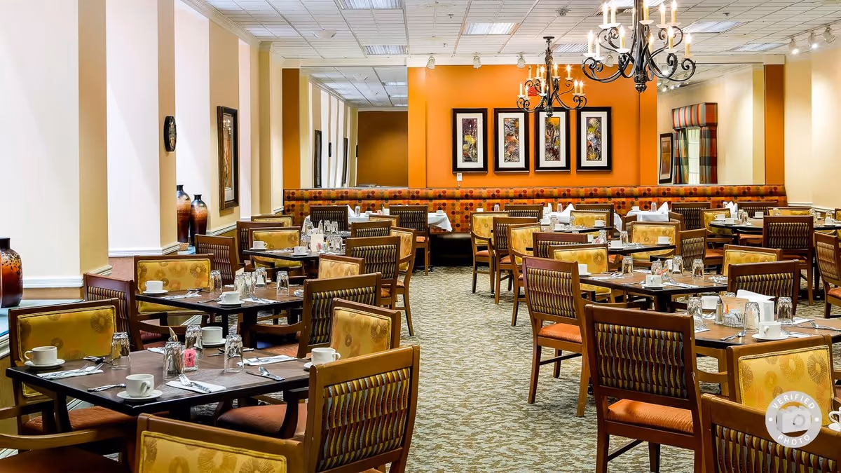 A spacious dining room with multiple tables set with cups, glasses, and silverware. The room features patterned carpet, cushioned chairs with yellow and orange upholstery, and an orange accent wall with framed artwork. Two chandeliers hang from the ceiling, and there is a long cushioned bench along the back wall.
