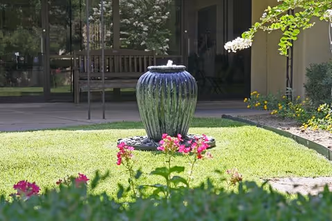 A green ceramic water fountain surrounded by pink flowers on a well-maintained lawn in an outdoor garden area. In the background, there is a wooden bench and glass doors leading to the interior of a building.