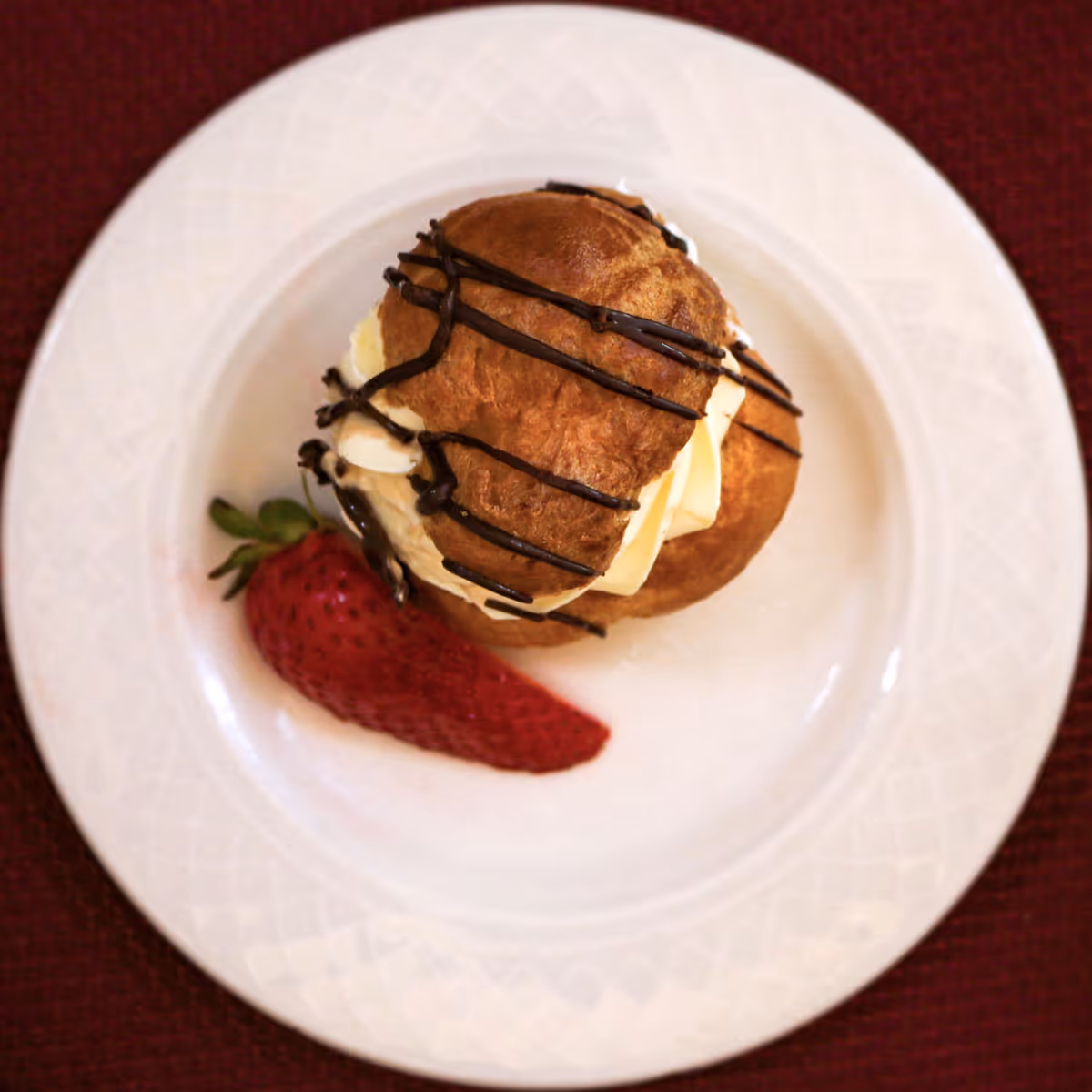 A cream puff drizzled with chocolate and a sliced strawberry on a white plate.