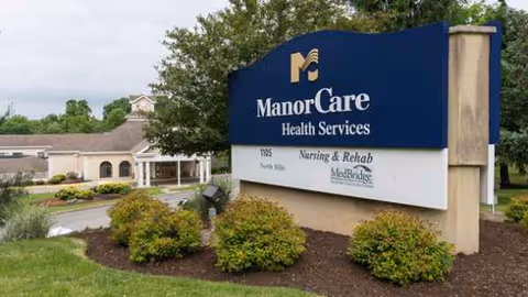 Exterior view of a blue ManorCare Health Services sign with landscaped surroundings.