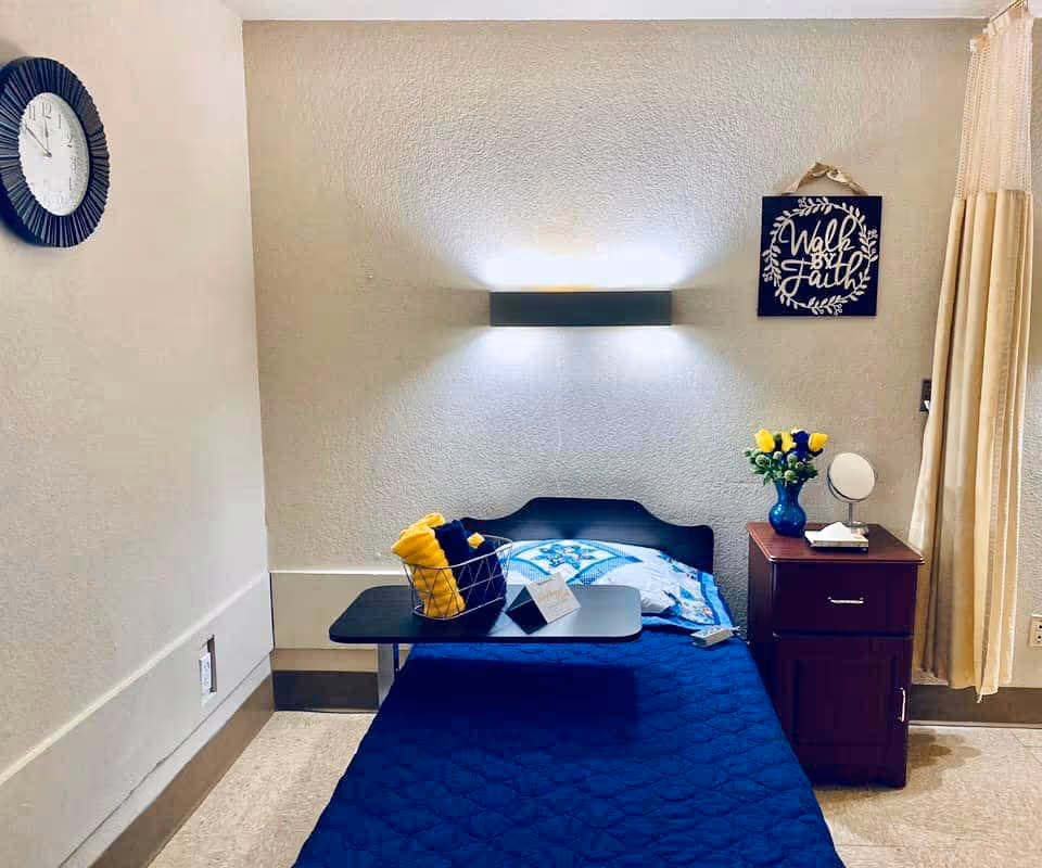 A small, neatly arranged bedroom in a senior living facility with a single bed covered in a blue quilt. A black overbed table holds a basket with yellow towels and a small card. On the right side, there is a wooden nightstand with a blue vase of yellow flowers, a small round mirror, and a tissue box. A wall light fixture illuminates the bed area, and a decorative sign reading 'Walk by Faith' hangs on the wall. A clock is mounted on the left wall, and beige curtains cover a window on the right.