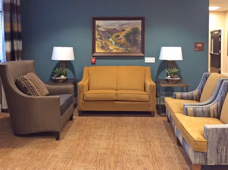 A cozy seating area in a senior living facility with a mustard yellow loveseat in the center, flanked by two side tables each with a lamp and a small plant. To the left is a brown armchair with a patterned cushion, and to the right is a striped sofa with mustard yellow cushions. A framed landscape painting hangs on the teal-colored wall behind the loveseat.