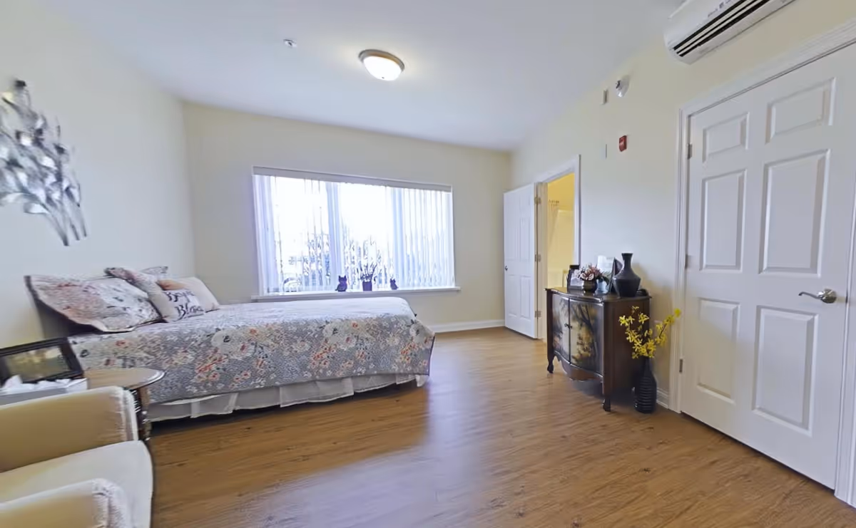 A bright bedroom with a single bed covered in a floral quilt, a beige armchair, a small round side table, a decorative wall piece, a wooden dresser with vases and flowers, and a large window with vertical blinds letting in natural light. The room has wooden flooring and white walls, with two white doors, one leading to a bathroom.