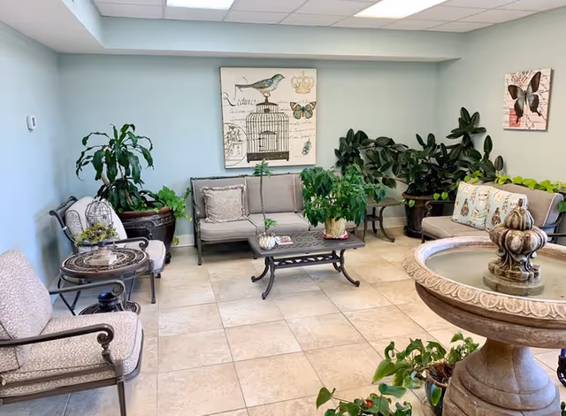 A cozy indoor seating area with cushioned chairs and a loveseat arranged around a small coffee table. The room features light blue walls adorned with butterfly and bird-themed artwork. Several large potted plants add greenery to the space, and a decorative stone water fountain is visible in the foreground.