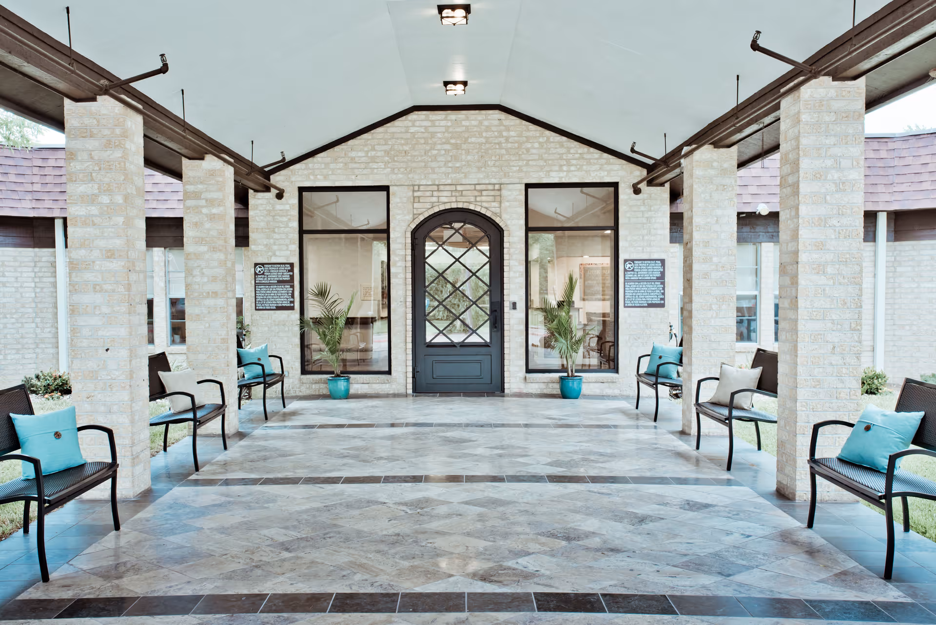 Covered outdoor seating area with stone pillars and tiled floor, featuring black metal benches with blue and beige cushions, potted plants near a black door with glass panels, and large windows on either side.